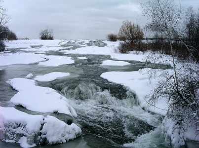 [A mini waterfall is surrounded by snow-covered ice in the portions of the river with less flow. Clumps of snow and flowing water is scattered over the image.]